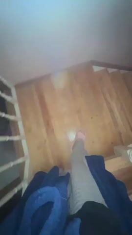 A person wearing a blue robe and grey pants is walking down a wooden staircase. The camera is pointed downwards, giving a first-person perspective. The stairs are well-lit.