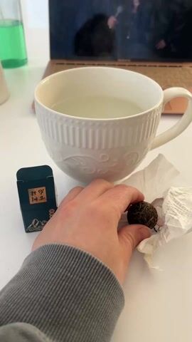 A person drops a small, dark, round object from a piece of crinkled white paper into a cup of clear liquid. The cup is white and ornate. A small black box with gold writing on it sits beside the cup. In the background, a laptop displays a video of a woman holding a microphone. A bottle with a green liquid is visible on the left. The surface is white. The person is wearing a gray long-sleeved shirt.