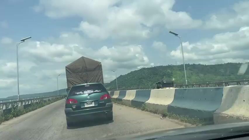The video captures a view from a moving vehicle on a bridge, likely in Adaha, Nigeria. The road is paved and appears to be a multi-lane highway. A green Toyota sedan is in the foreground, traveling on the road. Behind it, a large truck with a tarp-covered cargo is visible. Several other vehicles, including a truck and possibly more cars, are further down the road.

The setting is a bridge spanning a wide body of water, with lush green hills and mountains in the background. The sky is bright blue with scattered white clouds, suggesting a pleasant daytime, likely late morning or afternoon. The weather appears to be clear and sunny. Solar-powered streetlights are visible along the sides of the bridge.

The activity is simply traveling on the bridge. There are no specific events or actions depicted beyond vehicular movement. The mood conveyed is one of everyday travel and journeying through a scenic landscape.

Visible text includes:
- A license plate on the green Toyota, but the characters are not clearly legible.
- A sticker on the rear window of the Toyota, which appears to have the letters "WRC" and some other text, possibly related to a motorsport event.
- Signs on the concrete barriers lining the bridge, but the text is too small to read.
- A distant electrical pylon on the hillside.