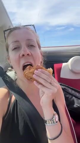 A woman in a black tank top is in the passenger seat of a car, eating a pastry. She is wearing a watch and several rings. The sky is visible through the open roof of the car, and the background shows car seats.