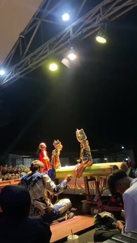 A group of people are watching a traditional puppet show outdoors at night. In the foreground, a puppeteer in ornate clothing is manipulating several puppets on a raised platform. The puppets appear to be made of carved leather or wood and are dressed in traditional attire. The stage is lit by spotlights, casting dramatic shadows. In the background, more puppets and spectators can be seen. The overall atmosphere is one of cultural performance and community gathering.