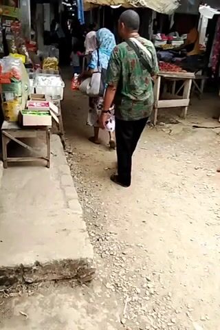 The video shows an outdoor market scene. In the foreground, a person wearing a green patterned shirt and black pants stands facing a stall filled with boxed goods. To the left, there is a concrete drainage ditch. In the background, another person wearing a blue headscarf and a patterned dress walks away from the camera. Other market stalls with various goods, including what appear to be red chili peppers, are visible further down the path. The ground is dirt and gravel.
