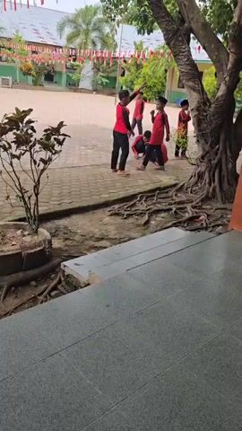 The video shows an outdoor scene, possibly a schoolyard. Several children, most wearing red shirts with black sleeves and black pants, are present. Two children are crouched down in the center, appearing to be engaged in an activity on the paved ground. One child to the left, also in red and black attire, is standing and appears to be gesturing. Another child in similar clothing stands to the right, with their arm raised. In the background, a person wearing a yellow garment and a headscarf walks away from the camera. Trees with exposed roots are visible in the foreground and midground. The ground is a mix of pavement and dirt. The scene appears to be during daylight.