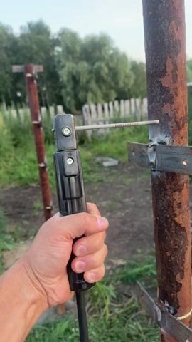 The video shows a close-up of a person's hand holding a welding electrode holder, which is clamped to a metal rod. The rod is attached to a rusty metal pole. There are sparks flying from the point of contact between the electrode and the pole, indicating that welding is in progress. The background is a blurry outdoor scene with greenery and a fence. The video is short, lasting only a few seconds, and captures the brief moment of welding.