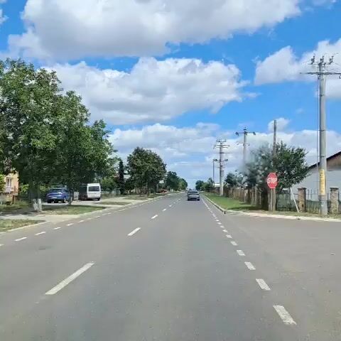The video shows a forward-facing view from a vehicle driving down a two-lane road under a blue sky with fluffy white clouds. Trees line the left side of the road, with houses and a white van visible behind them. A few cars are also present on the road, including one in the distance and others parked on the side. On the right side of the road, there are more trees, a fence, power lines with poles, and a stop sign. The road has clear white lane markings.