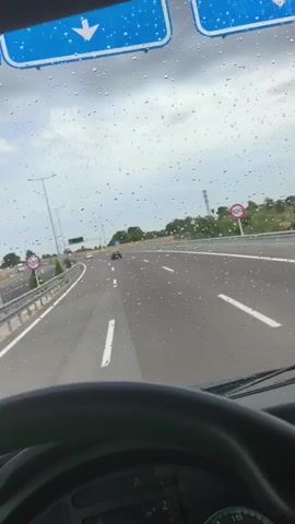 The video shows a view from inside a vehicle, looking out through the windshield. Raindrops are visible on the glass, distorting the view of the road ahead. The vehicle appears to be on a highway with multiple lanes. There are streetlights and signs visible alongside the road, and other vehicles can be seen in the distance. The sky is overcast with dark clouds. The interior of the vehicle is partially visible in the foreground, showing a steering wheel and dashboard. The video is short, lasting only a few seconds.