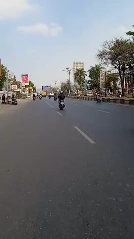 A video captures an overhead shot of a busy street in a city during the day. The camera pans slightly left to right, showcasing the road and the surrounding environment. Several people are seen riding scooters and motorcycles on the road. On the left side of the street, there are buildings, shops, and billboards. Palm trees and other greenery are visible on the right side, along with more buildings and streetlights. The sky is partly cloudy. The overall impression is of an active urban street scene.
