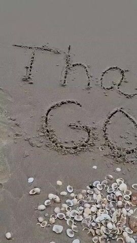 The image displays the words "Thank God" written in the sand on a beach. The sand is a light brown color and appears damp. The letters are formed by drawing lines into the surface of the sand, with some areas showing raised sand along the edges of the letters. Below the text, a pile of seashells is scattered on the sand. The seashells are of various sizes and shapes, predominantly white and beige, with some darker shells also visible. One larger, translucent object, possibly a piece of jellyfish or similar marine life, is situated within the pile of shells. The lighting suggests it is daytime, with soft shadows indicating overcast conditions or a time of day when the sun is not directly overhead. The overall scene evokes a sense of gratitude and peacefulness, commonly associated with a beach setting. The presence of the words "Thank God" written in the sand suggests a moment of reflection or appreciation. The location cues, such as the type of shells and the quality of the sand, are consistent with a tropical or subtropical beach environment.