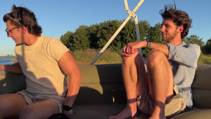 Two men are sitting in a vehicle, possibly a safari vehicle, with an olive-green padded seat, under a canopy. The man on the right is facing forward with his legs bent. He is wearing a light blue button-down shirt and tan shorts. He is smiling and looking towards something in front of him. The other man is only partially visible; we can see his arm and back. He is wearing a tan shirt and light-colored shorts. The vehicle is in an outdoor environment with a background of a river or lake and trees. The sky is blue. The video appears to have been taken during the day with good lighting conditions.