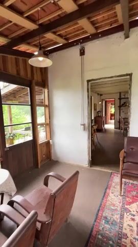 The video pans through a rustic, indoor space with wooden beams and a tiled roof. In the foreground, two wooden chairs and a small round table with a tablecloth are visible. Through glass doors and windows, lush green foliage and trees can be seen outside. The camera then moves into an adjacent room, revealing more wooden furniture, including tables and chairs, and shelves filled with books and decorative items. A sofa with a striped cushion sits against one wall, and a large wooden cabinet is positioned nearby. The overall impression is of a cozy, slightly dated, and well-lived-in interior with a strong connection to the outdoors.