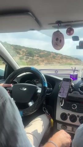 The video shows the interior of a car driving on a road. A person's hands are visible on the steering wheel and gear shift. The dashboard has a phone mounted with a navigation app open, a small dog figurine, an air freshener, and various other decorative items hanging from the rearview mirror. The landscape outside the car appears to be hilly and dry. The time stamp indicates the video is very short.