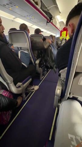 The video shows the interior of an airplane. Passengers are seated in rows, visible from the aisle. A person wearing a red jacket and neon yellow vest is walking up the aisle. The camera is positioned behind a seat, offering a view of the aisle and the passengers in the surrounding seats. A purple carpet runs down the aisle. The lighting is bright, and the interior appears clean.