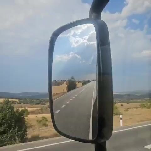 A side-view mirror of a vehicle reflects a rural road stretching into the distance under a bright, cloudy sky. The road has a white dashed line dividing the lanes and is flanked by dry, grassy fields and scattered trees. The sky is a vibrant blue with dramatic, sunlit clouds. The reflection shows the scenery passing by as the vehicle moves forward.