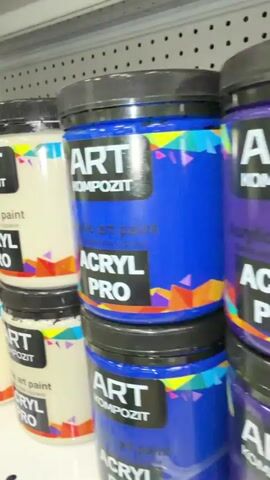 The video displays shelves stocked with jars of acrylic paint. The jars are predominantly cream-colored or brown, with black lids. They feature labels with the brand name "ART KOMPOZIT" and "ACRYL PRO." Further along the shelves, jars of a coppery metallic paint are visible, labeled "kompozit Metalliq ENAMEL ACRYLIC MAGIC EFFECT INTERIOR & EXTERIOR." The paint colors vary, showing cream, brown, and metallic copper. The environment appears to be a retail store or storage area, with the items neatly arranged on shelves. There are no people visible in the video, and no specific actions or interactions are depicted, other than the presentation of the products.