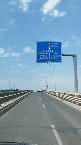 The video shows a view from a vehicle driving on a bridge. A blue road sign is visible at the beginning of the clip, indicating directions to various cities including Ciorogârla, Domnești, Constanța, Giurgiu, Alexandria, Ploiești, Târgoviște, and Pitești, with a roundabout symbol and a distance of 200 meters. The bridge has guardrails on both sides, and there are streetlights along the road. The sky is blue with scattered clouds. Several other vehicles are visible in the distance on the bridge. The camera is pointed forward, following the road as it curves. The latter part of the video shows the bridge continuing into the distance, with more vehicles and streetlights. The overall environment appears to be an urban or suburban setting.