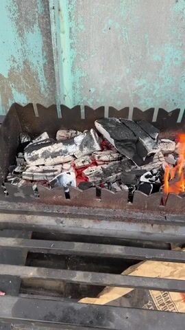 A close-up, top-down, slightly angled video captures burning charcoal in a grill. The charcoal is a mix of black, red, and white ash. Flames are visible on the right side of the grill. The grill is made of dark metal with a decorative, jagged edge on the top. The background consists of a weathered, teal-colored wall with patches of rust and dirt. Below the grill, a dark metal rack is visible, and a portion of a brown paper bag with Russian text is on the right.