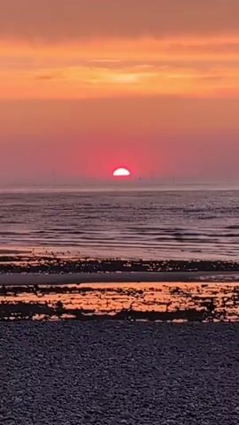 The video shows a sunset over the ocean. The sun is a bright red orb partially submerged below the horizon. The sky is filled with shades of orange and pink, with some clouds scattered across it. The ocean is calm, with gentle waves rolling towards the shore. The shore is covered in pebbles and has some wet sand visible. In the distance, there appear to be some wind turbines. There are no people or animals clearly visible in the video. The overall scene is peaceful and serene.