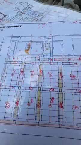 The video shows a construction site with scaffolding and building materials. The foreground features blueprints spread on the ground, with markings in red and yellow ink, suggesting project details or progress tracking. Behind the blueprints, stacks of metal scaffolding poles are visible. The background reveals a partially constructed building with more scaffolding and concrete structures. The scene is outdoors under a bright, cloudy sky, indicating daytime. There are no people visible in the frame.