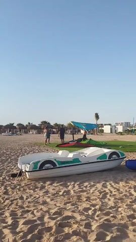 The video shows a beach on a clear day, with a white and green paddle boat parked on the sand in the foreground. In the background, three people stand near a blue canopy, and two windsurfing sails. There are palm trees, cabanas, and a building further down the beach. The people are walking around and interacting.