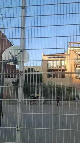 The video shows a fenced-in basketball court in an urban environment. Several people are on the court, some appear to be playing with a soccer ball. Buildings are visible in the background. The camera then moves along the outside of the fence to a sidewalk.