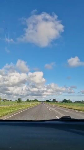 The video shows a view from inside a car driving on a paved road. The road has white lines marking the lanes. On either side of the road are fields of green grass and some trees. The sky is blue with white fluffy clouds. There are a few other cars visible in the distance on the road. The sun appears to be shining, creating bright lighting conditions. The dashboard of the car is visible at the bottom of the frame. The video appears to be taken on a sunny day, likely during daylight hours.