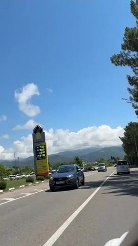 A roadside scene under a bright blue sky with scattered clouds. A tall, yellow and red gas price sign is visible on the left side of the frame. Several vehicles, including vans and cars, are parked along the side of the road or driving on it. The road is asphalt with white lane markings. Trees and mountains are visible in the background. The overall impression is a sunny day on a highway with a gas station.