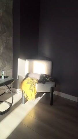 The scene is a corner of a room, illuminated by bright sunlight streaming in from the left. A white chair with silver legs sits against two dark gray walls. A yellow blanket with a pattern is draped over the chair. A gray cat is curled up on the chair, mostly obscured by the blanket. To the left of the chair is a small, round black table with a can of deodorant on it. The floor is light-colored wood. Someone is making a "tsk tsk" sound in the background.