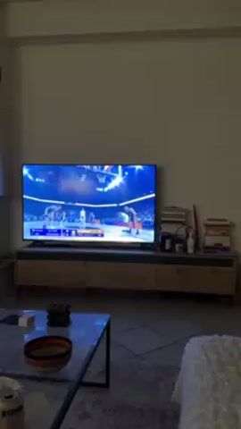 The video shows an indoor shot of a living room. In the background, a television displays a basketball game. The TV is placed on a long, low wooden cabinet, and several books are stacked on the right side of the cabinet. In the foreground, there is a glass-topped coffee table with a few decorative items. The camera focuses on a person petting a cat, which is mostly white with some brown markings. The cat's eyes are closed as it is being petted.