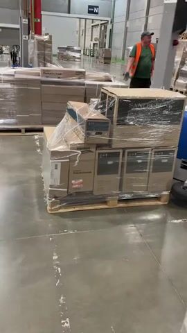 A man in an orange safety vest and a cap is walking in a warehouse. In front of him, a pallet of boxes wrapped in plastic is being moved by a blue and gray machine with a wheel. The machine has a control panel with buttons and a small screen. The pallet is on a wooden base. Other pallets of boxes are visible in the background, along with warehouse shelving and industrial lighting. The floor is concrete.
