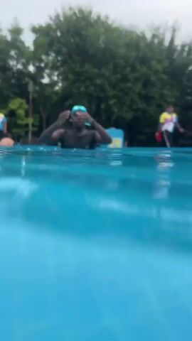 An underwater shot of a person swimming in a pool. The person is wearing goggles and a dark swimsuit. The water is blue and clear, with sunlight filtering from above. The camera is moving, giving a sense of motion and depth. The person's face is partially obscured by water and movement.