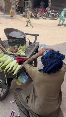 The video shows a man seated on the ground next to a pushcart, holding a water bottle to his head. The man is wearing a brown shalwar kameez and a blue checkered scarf around his head. The pushcart is loaded with corn on the cob, some pieces of wood, and a metal cooking pot filled with dark-colored material, possibly sand. A red cloth and a small dish sit among the corn. In the background, a boy walks past, carrying a bag. There are parked motorcycles, a white car, and buildings lining the street. The ground is dusty and appears to be a roadside.
