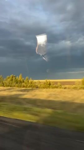 This is a video of a landscape seen through a moving vehicle, likely a car. The perspective is from inside the vehicle looking out through the window. The foreground is blurred, suggesting motion. The middle ground shows a strip of trees with green and yellow foliage, bordering a field of dry, golden grass. Beyond that, a darker, more distant field is visible under a cloudy sky. A prominent reflection on the window obscures part of the view, showing a distorted image of a person or object and possibly the interior of the vehicle, as well as the sky. The sky itself is dramatic, with dark, heavy clouds dominating the upper portion of the frame, interspersed with patches of lighter sky and sunlight. The overall impression is of a transitional weather event, possibly a storm approaching or receding, viewed from a moving perspective.