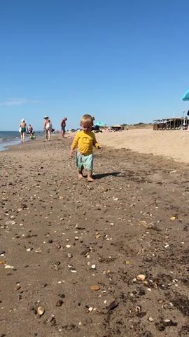A young child with blonde hair, wearing a yellow long-sleeved shirt with a lion graphic and light blue shorts with a fish pattern, walks towards the camera on a sandy beach. The child's face is slightly dirty with sand. In the background, the ocean is visible on the left, with several people walking along the shoreline. Beach umbrellas and structures are present further down the beach on the right. The sky is clear and blue.