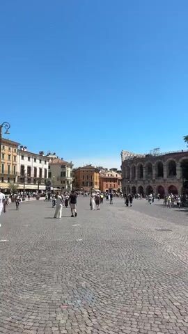 A vertical, wide shot captures an outdoor public square paved with cobblestones under a clear blue sky. The square is populated by a moderate number of people walking in various directions. On the left side of the frame, buildings with balconies and awnings line the square, with tables and chairs from what appears to be a cafe visible. In the background, on the right side of the frame, a large, ancient Roman amphitheater with arched entrances is partially visible. The sun is shining, casting shadows on the ground, suggesting it is daytime. The overall atmosphere appears to be that of a tourist destination or a bustling city center.