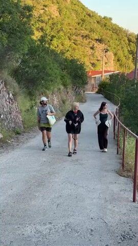 The video shows three people walking on a paved path next to a hillside covered in trees. The path curves to the right and has a metal railing on the right side. A man in a gray t-shirt, black shorts, and a white hat walks ahead, carrying a light-colored bag over his shoulder. Behind him, two women walk together. One, in a black hoodie and shorts, is looking at her phone. The other, in a dark tank top and brown pants, walks beside her. Houses with red roofs are visible on the hillside in the background. The sun is shining, casting shadows and illuminating the green foliage.