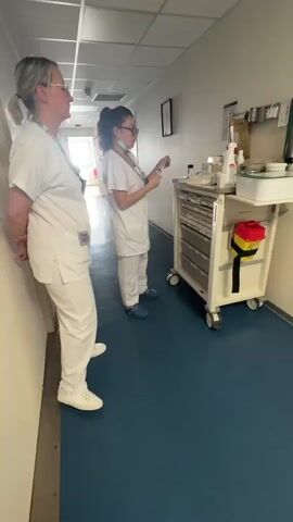 Two women in medical scrubs are in a hospital or clinic hallway. The woman on the left, with blonde hair tied back, is wearing glasses, a white scrub top and pants, and white sneakers, and is looking straight ahead. The woman on the right, with dark hair in a ponytail, glasses, and a light blue face mask, is also wearing a white scrub top and pants, and blue clogs. She is holding what appears to be a syringe or medical device and is standing next to a white medical cart with multiple drawers. A red and yellow sharps container with a biohazard symbol is attached to the side of the cart. Various medical supplies are visible on top of the cart. The hallway has a blue floor and a window at the far end, providing natural light. Both women are wearing lanyards with ID badges. The woman on the right appears to be preparing medication or supplies, while the woman on the left observes.
