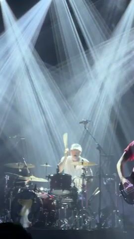 The video shows a drummer performing onstage, bathed in white and then yellow spotlight. He wears a white shirt, light pants, and a white baseball cap. He is behind a drum kit. To the right of the frame, a person in a pink shirt plays a bass guitar. The background is dark with bright, converging spotlights.