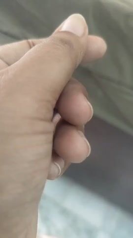 A close-up shot focuses on a person's left hand, with fingers curled to form a heart shape. The thumb and index finger are extended, with their tips touching to create the top curves of the heart, while the middle three fingers are gently bent. The hand is positioned against a blurred background that appears to be a textured fabric in muted colors, possibly a blanket or upholstery. The lighting is soft, highlighting the contours of the hand and the subtle details of the skin.