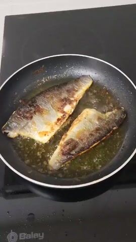 The video shows two fish fillets frying in a pan with oil on a black Balay induction cooktop. The fish skin side is browned, and the oil is bubbling. Steam rises from the pan. The countertop and wall behind the stove are white.  The camera is static and close up.