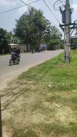 The video shows a roadside scene with a paved road curving to the right. On the left side of the road, there are buildings and trees. A single motorcycle with a rider is visible on the road, moving away from the camera. On the right side of the road, there is a grassy embankment, a utility pole with electrical equipment and wires, and more trees. The sky is visible overhead, appearing partly cloudy. The overall impression is a rural or semi-rural roadside environment on a sunny day.