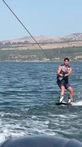 Here is a concise description of the video:

A man wakeboards on a body of water on a sunny day. He is wearing a life vest, shorts, and wakeboarding boots, holding onto a rope that pulls him across the water. In the background, there is land with some vegetation and low buildings. The water is choppy, and the sky is clear.