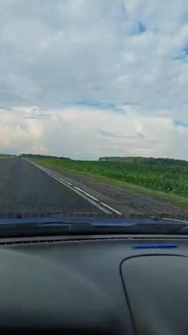 This is a first-person view from inside a car, looking out the windshield. The car is driving on an asphalt road, which has two white lines marking the lanes. The road stretches into the distance, disappearing over a gentle hill. To the right of the road is a wide field of green crops, possibly corn or sunflowers, with a line of trees in the far distance. The sky is overcast with large, white clouds and patches of blue sky. The dashboard of the car is visible in the foreground, dark gray in color. There is a hint of a blue object on the dashboard. The video appears to be taken during daylight.