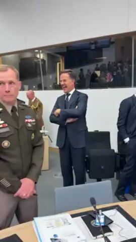 Here is a description of the video:

The video shows Dutch Prime Minister Mark Rutte standing in a room with other officials. He is wearing a dark suit, and he is smiling and laughing. He shakes hands with another man wearing a light blue suit. The other officials in the room are wearing military uniforms and business suits. The room is well-lit and has a large window. There is a table in the foreground with papers and a microphone on it.