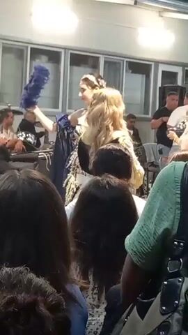 A group of people are gathered outdoors, likely for a performance or event. In the foreground, the backs of several audience members are visible, with one person wearing a green shirt and carrying a black bag. The middle ground features performers in traditional or costume attire. A woman in a blue and gold ornate dress with a blue feather fan is on the left. To her right, a woman in a purple vest over a white and gold dress is holding a silver tray. Behind her, a band is set up with drums and electronic equipment. Further to the right, a young girl in a yellow dress holds a white fan. The background shows a building with windows and overhead lighting. The overall atmosphere appears festive and engaged.