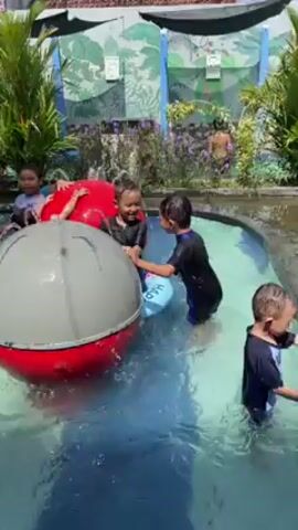 Several children swim and play in a shallow outdoor pool on a sunny day. A set of red and grey bars with a spout is in the center of the pool, spraying water. A small slide is in the background. There are several adults, presumably supervising, dressed in modest clothing. Some children play with plastic flotation devices. A covering shades the pool.
