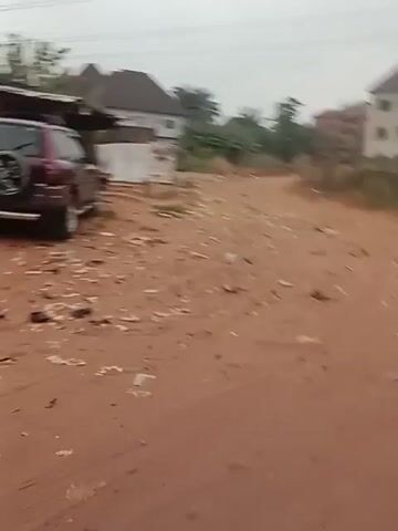 The video shows a landscape featuring a maroon SUV parked on a dirt lot. The ground is a reddish-brown color, and there are small pieces of debris scattered around. In the background, there are several buildings, power lines, and trees. The sky is overcast, creating a muted, grey tone. The camera pans across the scene, starting with a close-up of the SUV and then widening to show more of the surrounding area.