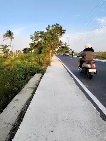 A video shows a sidewalk next to a road in a rural area. The sidewalk is made of concrete and has a gutter next to it. The road is paved and has a white line marking the edge. There are trees and vegetation on the side of the road. In the distance, there are power lines and a few buildings. Several people on motorcycles are seen riding on the road. The sky is clear and blue.