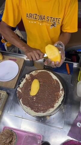 A person wearing an Arizona State T-shirt and clear plastic gloves is making a crepe. They are using a knife to slice a mango and place the slices on top of a crepe covered in chocolate spread. The crepe is on a heated metal surface. There is a stack of toothpicks in a pink container to the side. Other items are also on the cart.