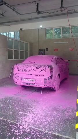 The video shows a pink foam-covered car at a car wash. A person, wearing a blue shirt and dark shorts, is visible in the background on the right, holding a hose and spraying the car. The car is positioned on a grate within the car wash bay, and the floor is covered in pink foam and water. The car wash bay appears to be an indoor facility with concrete walls, a high ceiling, and fluorescent lighting. Windows with blinds are visible in the background, and there are several red warning signs on the walls.