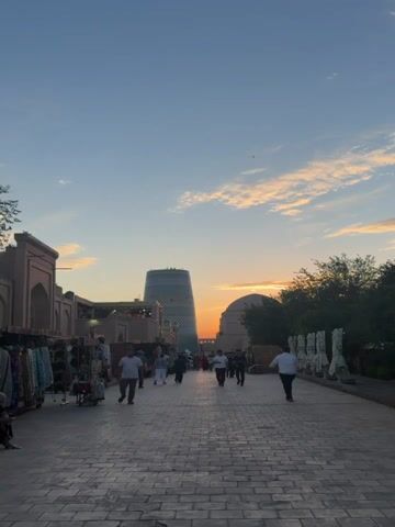 The video captures a scene at dusk in an outdoor marketplace. A paved walkway, constructed with light-colored bricks, stretches into the distance. On the left side of the walkway, market stalls are set up with merchandise displayed on racks, featuring various textiles and clothing items. Several people are walking along the pathway, some individually, others in small groups. In the background, distinctive architectural structures are visible: a tall, cylindrical building with a patterned facade and a large, rounded dome. The sky above is a gradient of blue and orange, with scattered clouds illuminated by the setting sun, creating a warm, atmospheric glow. Trees are present on the right side of the image, adding to the outdoor setting. The overall ambiance suggests a lively yet tranquil evening in a historical or cultural location.